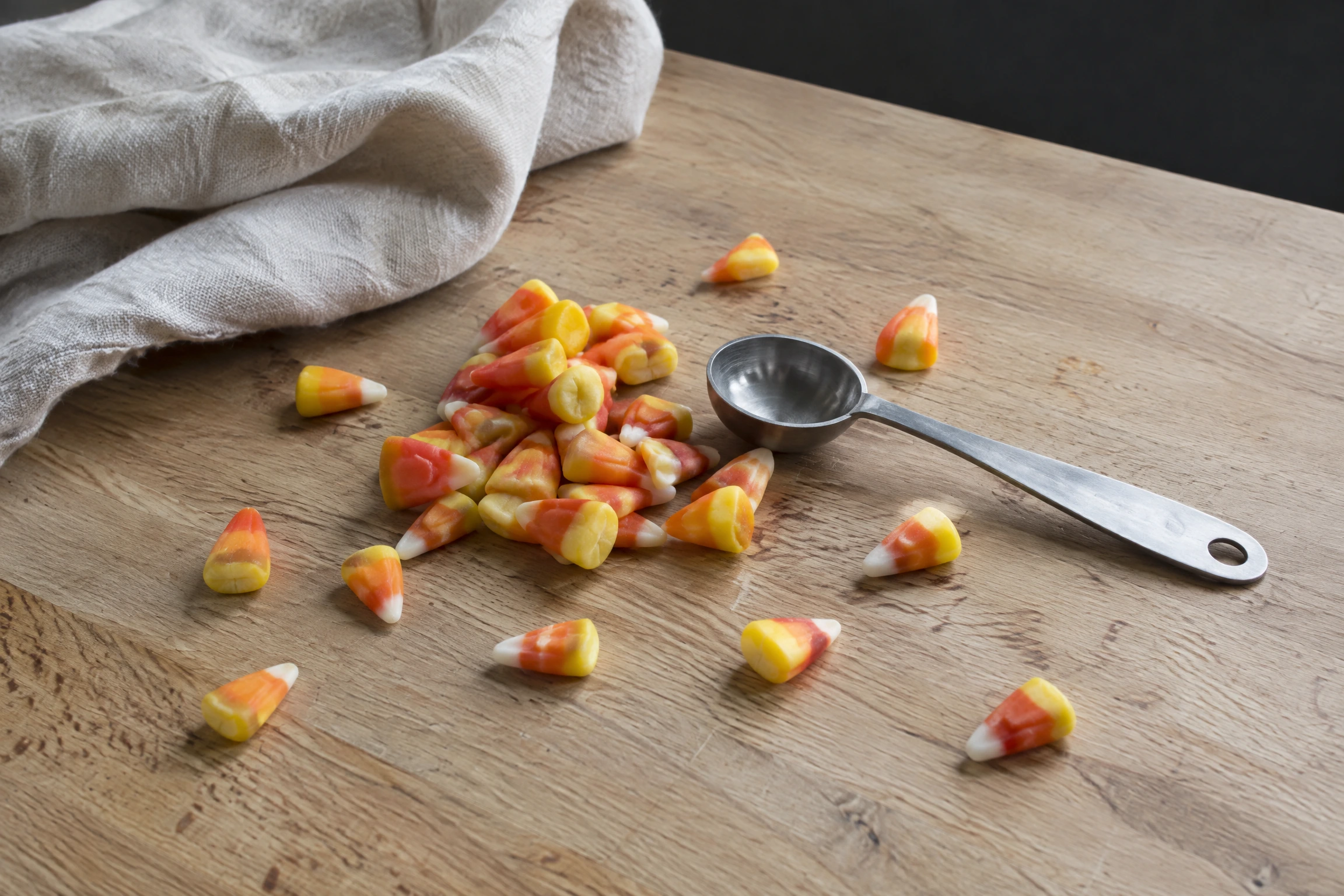 Des bonbons candy corn jaunes, oranges et blancs éparpillés et en petit tas sur un plan de travail en bois clair, avec une cuillère en métal posée à côté.