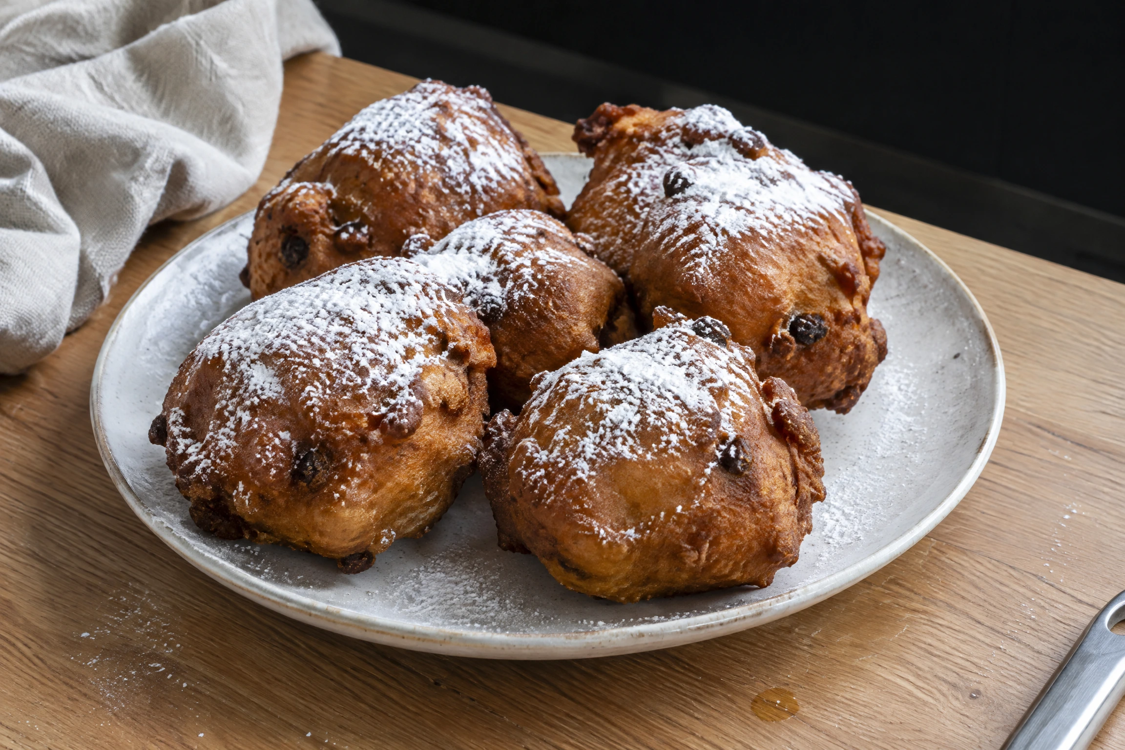 Assiette de beignets dorés saupoudrés de sucre blanc, posés sur un plan de travail en bois clair.